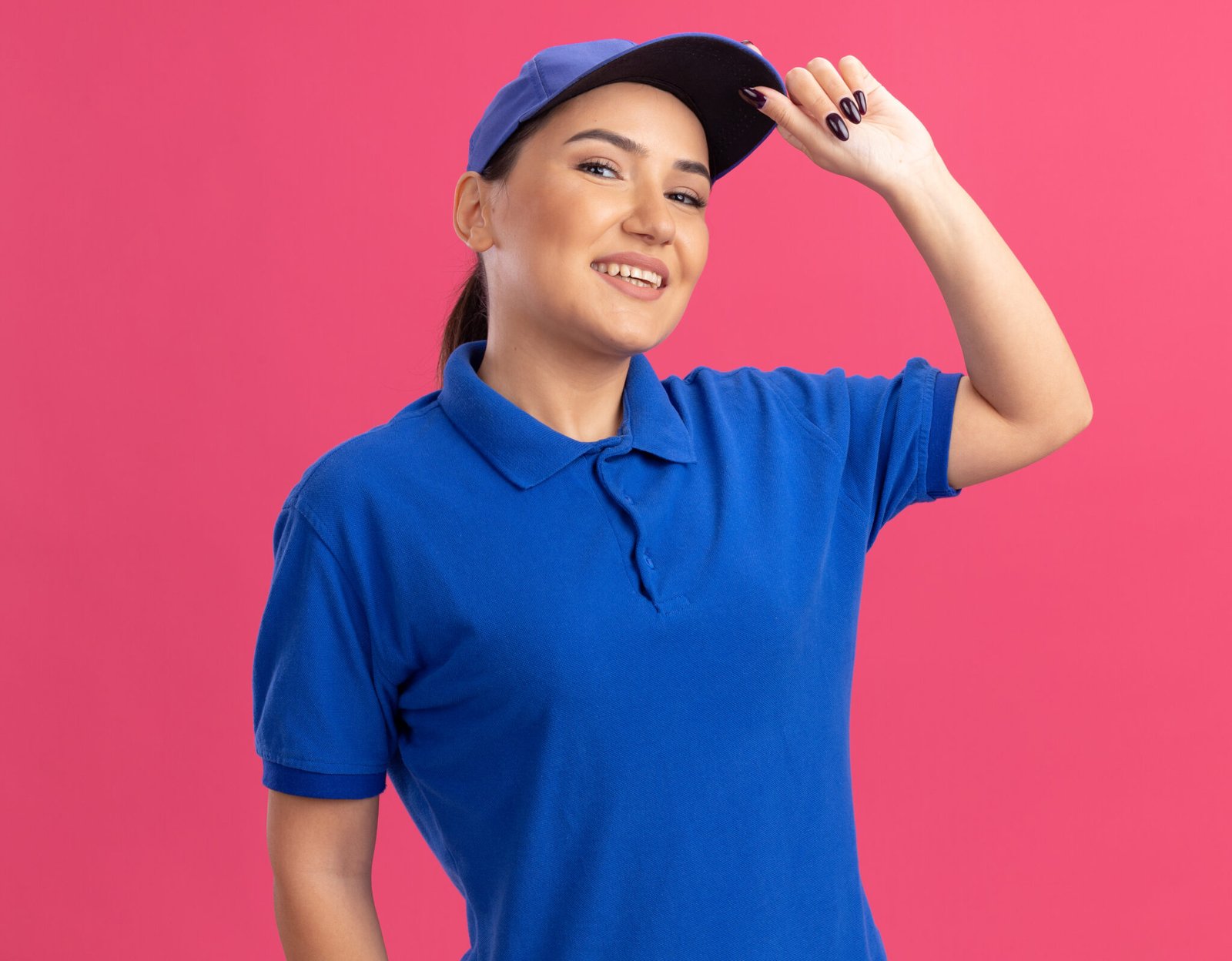 young delivery woman in blue uniform and cap looking at camera smiling confident touching her cap standing over pink background