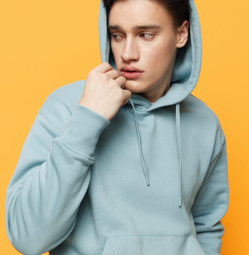 a handsome teenager stands on an orange background in a light hoodie and adjusts the hood with his hands looking to the side. High quality photo
